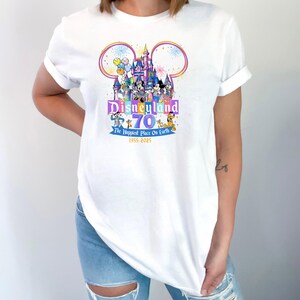 May include: White t-shirt featuring a colorful Disneyland 70th anniversary graphic. The design includes a castle, Mickey Mouse ears, and various Disney characters. Text reads "Disneyland 70 The Happiest Place On Earth 1955-2025".