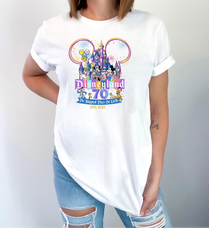 May include: White t-shirt featuring a colorful Disneyland 70th anniversary graphic. The design includes a castle, Mickey Mouse ears, and various Disney characters. Text reads "Disneyland 70 The Happiest Place On Earth 1955-2025".