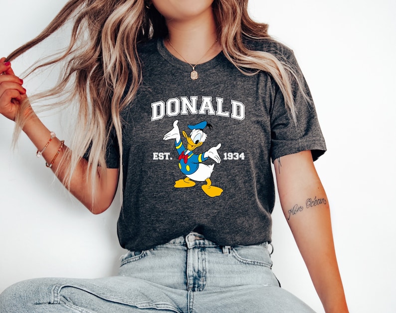 May include: A dark grey t-shirt with a white graphic of Donald Duck. The graphic includes the text "DONALD EST. 1934".