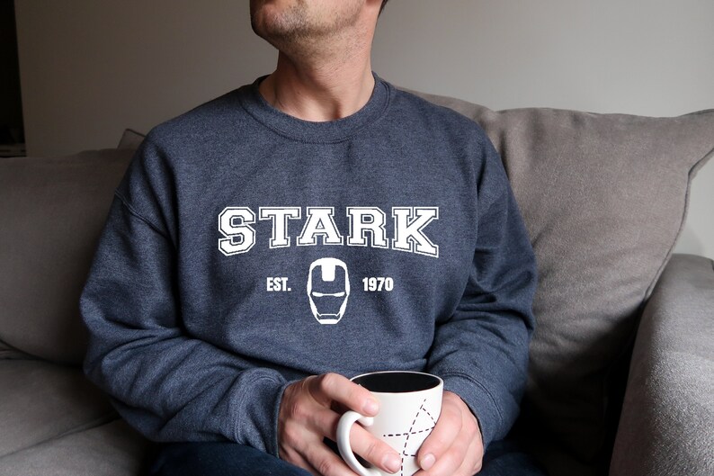 Pode incluir: Um moletom azul marinho com a palavra "STARK" em letras brancas, juntamente com "EST. 1970" e um gr&aacute;fico do capacete do Homem de Ferro. A pessoa est&aacute; segurando uma caneca branca com interior preto e design geom&eacute;trico.