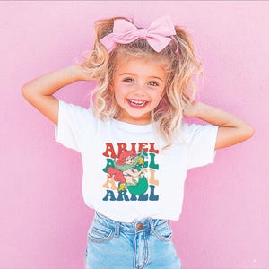 May include: A white t-shirt featuring a colourful graphic of a mermaid with the text "Ariel" repeated in a stacked pattern.