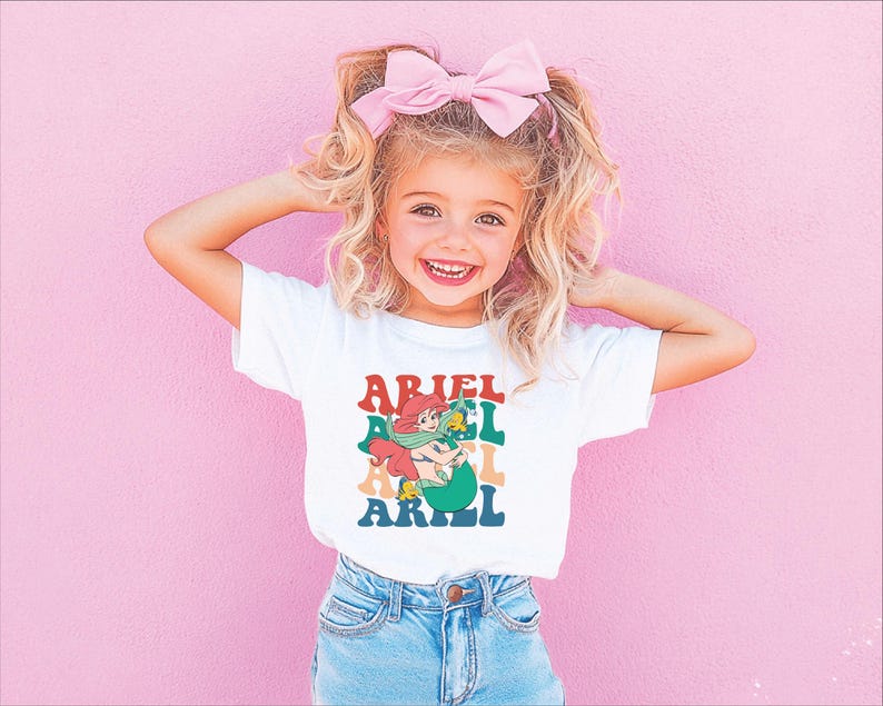 May include: A white t-shirt featuring a colourful graphic of a mermaid with the text "Ariel" repeated in a stacked pattern.