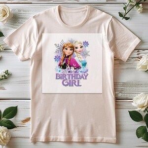 K&ouml;nnte beinhalten: Hellrosa T-Shirt mit einem Aufdruck von zwei Zeichentrickm&auml;dchen und dem Text "BIRTHDAY GIRL" in Lila. Die Grafik enth&auml;lt Schneeflocken und florale Elemente. Das Shirt wird auf einer hellen Holzoberfl&auml;che mit wei&szlig;en Rosen und Gr&uuml;nzeug pr&auml;sentiert.