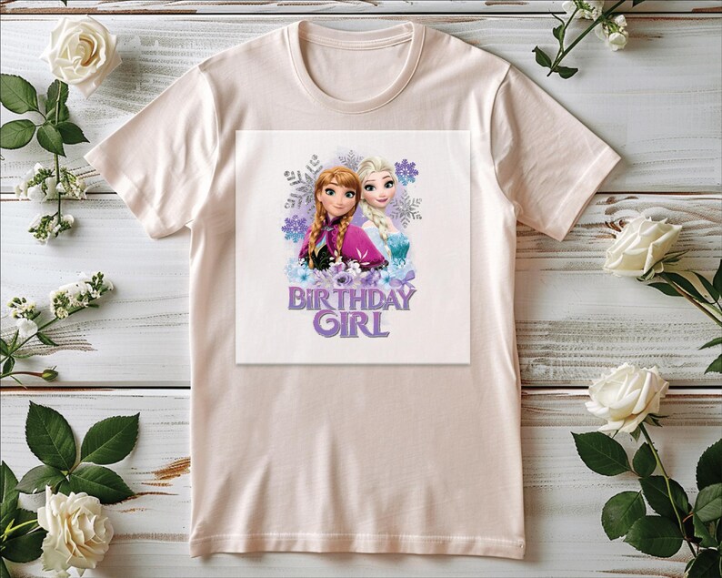 K&ouml;nnte beinhalten: Hellrosa T-Shirt mit einem Aufdruck von zwei Zeichentrickm&auml;dchen und dem Text "BIRTHDAY GIRL" in Lila. Die Grafik enth&auml;lt Schneeflocken und florale Elemente. Das Shirt wird auf einer hellen Holzoberfl&auml;che mit wei&szlig;en Rosen und Gr&uuml;nzeug pr&auml;sentiert.