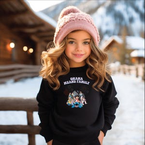 K&ouml;nnte beinhalten: Ein schwarzes Sweatshirt mit dem Text "OHANA MEANS FAMILY" und einer Disney-Figurengrafik. Das Sweatshirt wird von einer jungen Person getragen, die auch eine rosa Strickm&uuml;tze tr&auml;gt. Der Hintergrund zeigt eine verschneite Landschaft.
