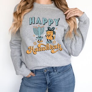 May include: A gray sweatshirt with the words "HAPPY Hanukkah" in a retro font. The design features a cartoon menorah and a gift box with arms and legs. The menorah has lit candles, and the gift box has a blue bow.