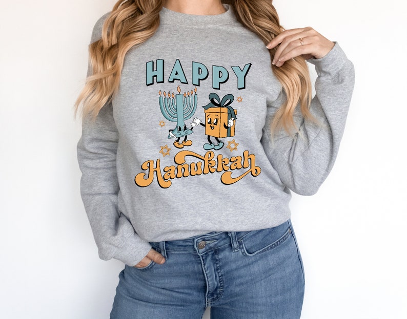 May include: A gray sweatshirt with the words "HAPPY Hanukkah" in a retro font. The design features a cartoon menorah and a gift box with arms and legs. The menorah has lit candles, and the gift box has a blue bow.