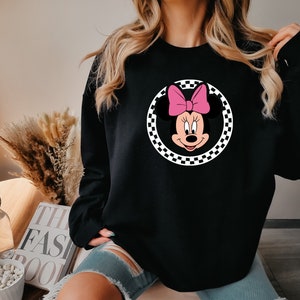 Puede incluir: Sudadera negra con un gr&aacute;fico circular a cuadros que presenta una imagen de dibujos animados de Minnie Mouse con un lazo rosa.