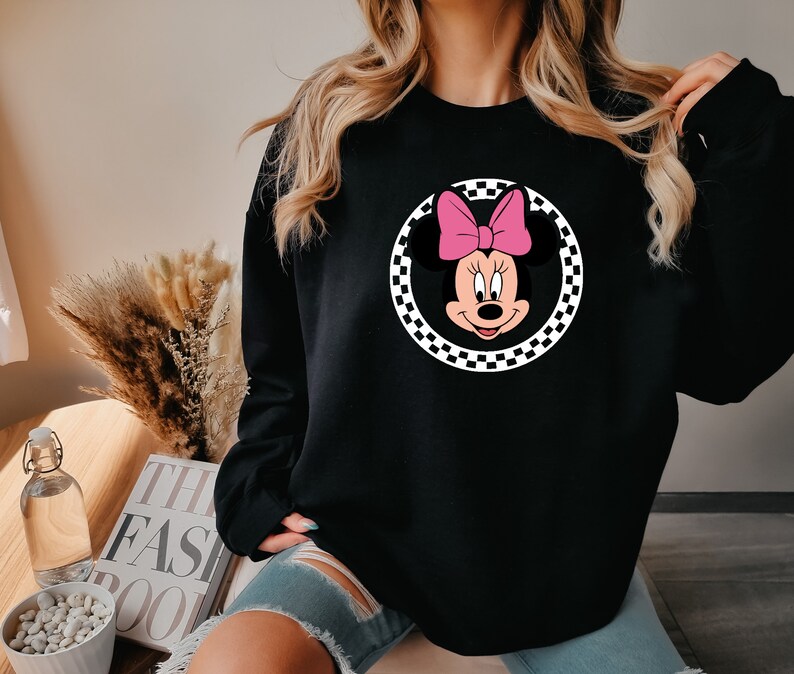Puede incluir: Sudadera negra con un gr&aacute;fico circular a cuadros que presenta una imagen de dibujos animados de Minnie Mouse con un lazo rosa.