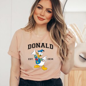 May include: A peach-colored t-shirt with a cartoon image of Donald Duck. The shirt has black text that reads "DONALD EST. 1934".