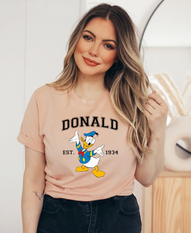 May include: A peach-colored t-shirt with a cartoon image of Donald Duck. The shirt has black text that reads "DONALD EST. 1934".