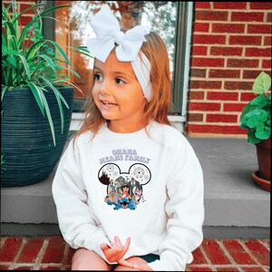 K&ouml;nnte beinhalten: Ein wei&szlig;es Sweatshirt mit dem Text "Ohana Means Family" in Lila, mit einer Cartoon-Grafik von Charakteren. Das Sweatshirt wird von einem kleinen Kind mit einem wei&szlig;en Stirnband und einer Schleife getragen.