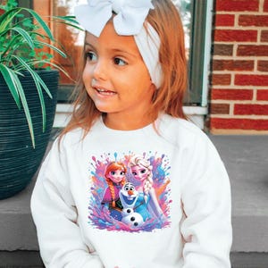 May include: A young girl wearing a white sweatshirt featuring a colorful graphic of Anna, Elsa, and Olaf from the movie Frozen. She has a white headband and a large white bow in her hair. The background includes a brick wall and greenery.