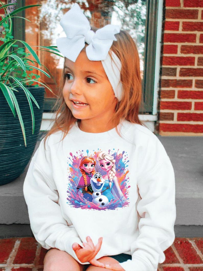 May include: A young girl wearing a white sweatshirt featuring a colorful graphic of Anna, Elsa, and Olaf from the movie Frozen. She has a white headband and a large white bow in her hair. The background includes a brick wall and greenery.