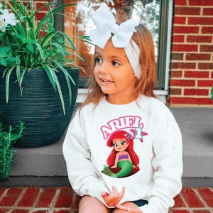 Może przedstawiać: Mała dziewczynka w białej bluzie z nadrukiem z kresk&oacute;wki Arielki. Bluza ma napis "ARIEL" w r&oacute;żowych literach. Ma na sobie białą opaskę z kokardą. Tło obejmuje rośliny doniczkowe i ceglaną ścianę.