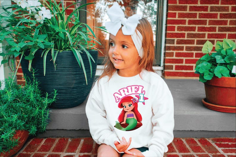 Może przedstawiać: Mała dziewczynka w białej bluzie z nadrukiem z kresk&oacute;wki Arielki. Bluza ma napis "ARIEL" w r&oacute;żowych literach. Ma na sobie białą opaskę z kokardą. Tło obejmuje rośliny doniczkowe i ceglaną ścianę.