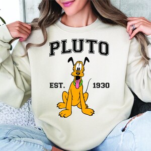 Puede incluir: Una sudadera de color crema con una imagen de dibujos animados de Pluto el perro. El texto "PLUTO" est&aacute; impreso en negro con un contorno blanco. El texto "EST. 1930" est&aacute; impreso debajo de la imagen.