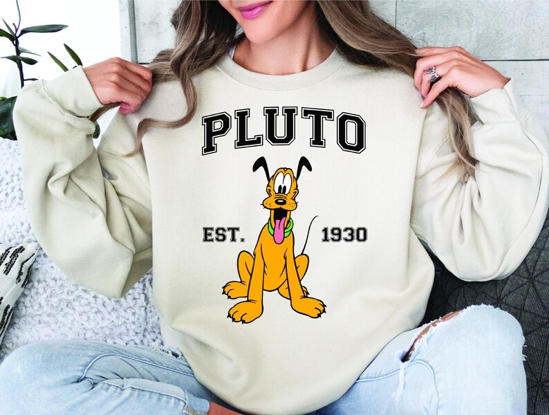 Puede incluir: Una sudadera de color crema con una imagen de dibujos animados de Pluto el perro. El texto "PLUTO" est&aacute; impreso en negro con un contorno blanco. El texto "EST. 1930" est&aacute; impreso debajo de la imagen.