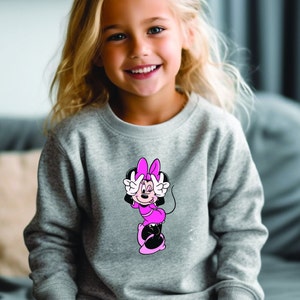 Może przedstawiać: Szara bluza z nadrukiem Myszki Minnie w kolorze r&oacute;żowym i czarnym. Postać z kresk&oacute;wki robi znak pokoju. Bluza jest noszona przez małe dziecko z blond włosami.