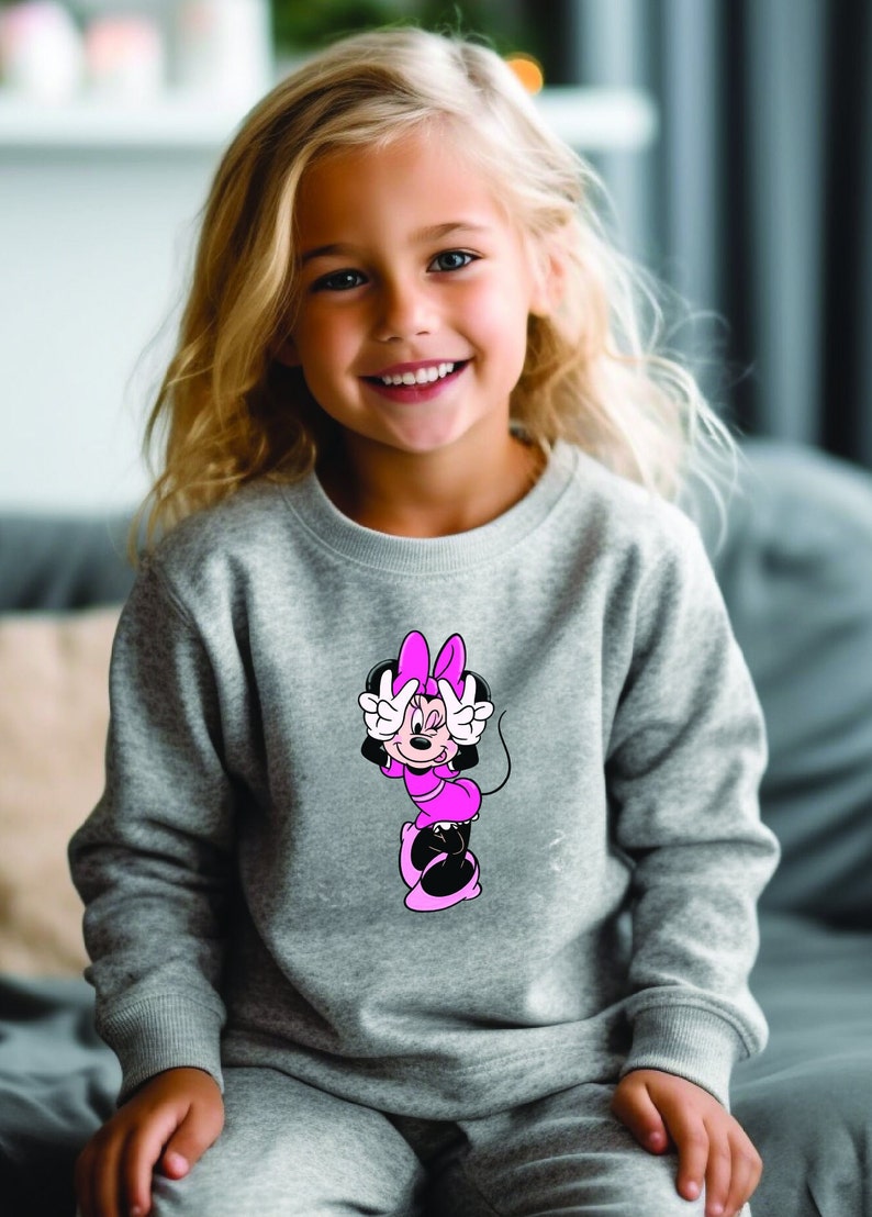 Może przedstawiać: Szara bluza z nadrukiem Myszki Minnie w kolorze r&oacute;żowym i czarnym. Postać z kresk&oacute;wki robi znak pokoju. Bluza jest noszona przez małe dziecko z blond włosami.