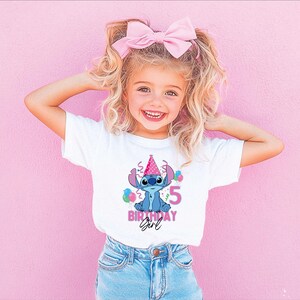 May include: A young girl wearing a white t-shirt featuring a Stitch graphic, a pink birthday hat, and the number 5. The shirt also says "Birthday Girl" in pink. She is wearing blue jeans and a pink bow in her hair.