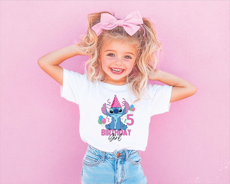 May include: A young girl wearing a white t-shirt featuring a Stitch graphic, a pink birthday hat, and the number 5. The shirt also says "Birthday Girl" in pink. She is wearing blue jeans and a pink bow in her hair.