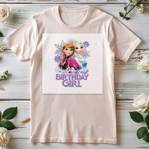 K&ouml;nnte beinhalten: Hellrosa T-Shirt mit einem Aufdruck von zwei Zeichentrickm&auml;dchen und dem Text "BIRTHDAY GIRL" in Lila. Die Grafik enth&auml;lt Schneeflocken und florale Elemente. Das Shirt wird auf einer hellen Holzoberfl&auml;che mit wei&szlig;en Rosen und Gr&uuml;nzeug pr&auml;sentiert.