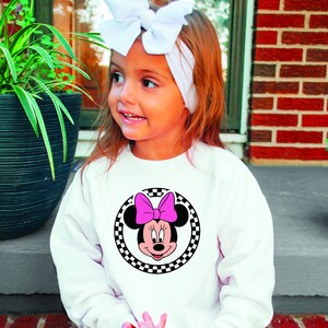 Puede incluir: Una sudadera blanca con un c&iacute;rculo de cuadros negros y blancos con una imagen de dibujos animados de Minnie Mouse con un lazo rosa.