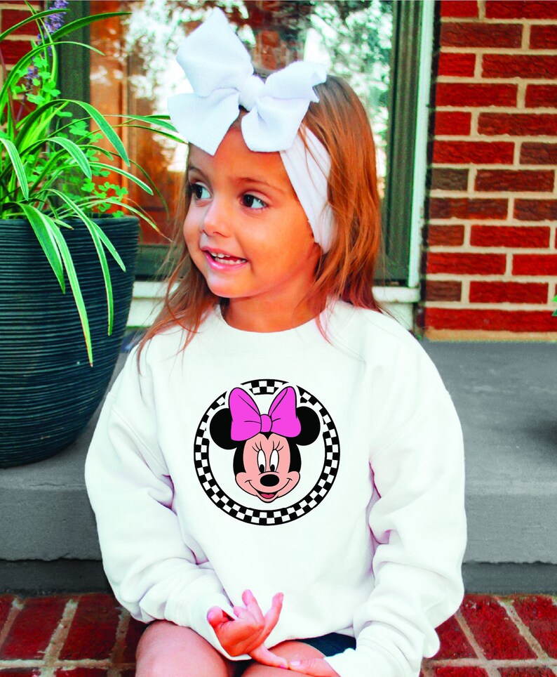 Puede incluir: Una sudadera blanca con un c&iacute;rculo de cuadros negros y blancos con una imagen de dibujos animados de Minnie Mouse con un lazo rosa.