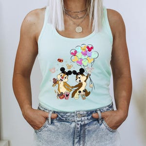 May include: A light blue tank top featuring a colorful graphic of Chip and Dale holding hands and balloons. The cartoon characters are brown with black ears and are surrounded by food and balloons. The tank top is worn with denim shorts.