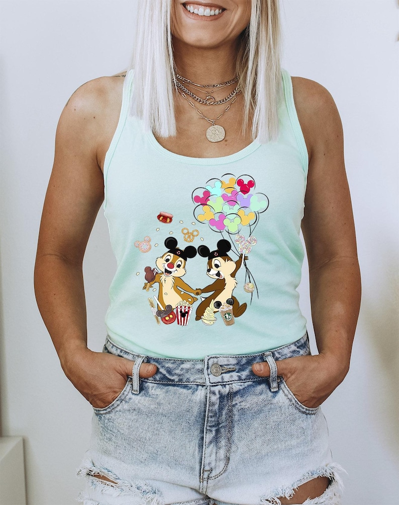 May include: A light blue tank top featuring a colorful graphic of Chip and Dale holding hands and balloons. The cartoon characters are brown with black ears and are surrounded by food and balloons. The tank top is worn with denim shorts.