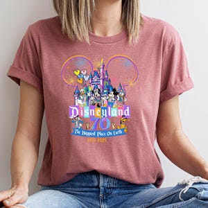 May include: A maroon t-shirt featuring a colorful Disneyland graphic. The design includes the Disneyland castle, Mickey Mouse ears, and various Disney characters. Text reads "Disneyland 70 The Happiest Place On Earth 1955-2025".