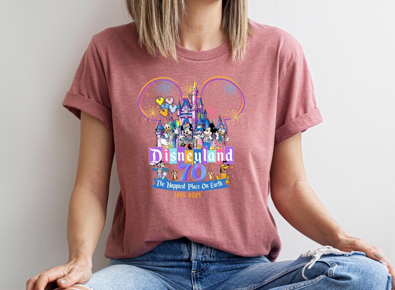 May include: A maroon t-shirt featuring a colorful Disneyland graphic. The design includes the Disneyland castle, Mickey Mouse ears, and various Disney characters. Text reads "Disneyland 70 The Happiest Place On Earth 1955-2025".
