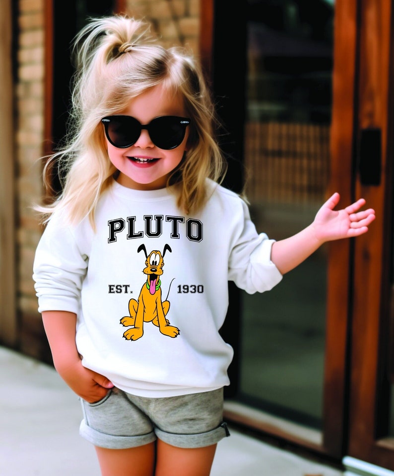 Puede incluir: Una sudadera blanca con una imagen de dibujos animados de Pluto el perro. La sudadera tiene el texto "PLUTO" en letras negras sobre la imagen y "EST. 1930" en letras negras debajo de la imagen.