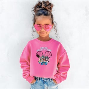 K&ouml;nnte beinhalten: Ein leuchtend pinkfarbenes Sweatshirt mit dem Text "OHANA MEANS FAMILY" und einer Grafik von Disney-Figuren. Das lang&auml;rmlige Sweatshirt wird von einer jungen Person getragen, die herzf&ouml;rmige Sonnenbrillen und blaue Jeans tr&auml;gt.