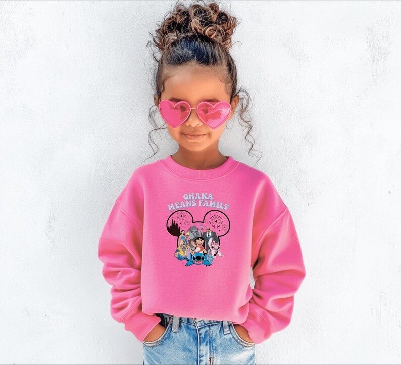 K&ouml;nnte beinhalten: Ein leuchtend pinkfarbenes Sweatshirt mit dem Text "OHANA MEANS FAMILY" und einer Grafik von Disney-Figuren. Das lang&auml;rmlige Sweatshirt wird von einer jungen Person getragen, die herzf&ouml;rmige Sonnenbrillen und blaue Jeans tr&auml;gt.