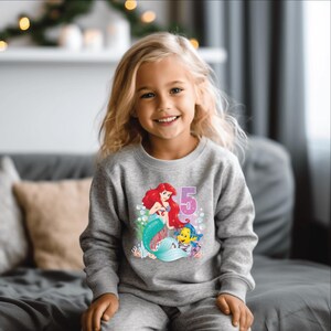 May include: A gray sweatshirt with a graphic of Ariel the Little Mermaid and Flounder. The graphic also includes the number 5.