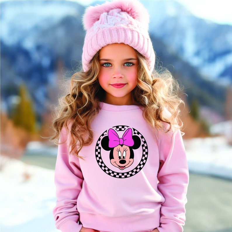 Puede incluir: Una sudadera rosa con un c&iacute;rculo de cuadros negros y blancos que presenta una imagen de dibujos animados de Minnie Mouse con un lazo rosa. La sudadera tiene mangas largas y cuello redondo.