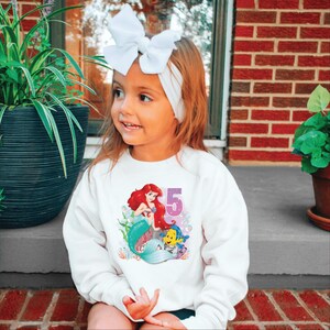 May include: White sweatshirt with a graphic of Ariel the Little Mermaid and Flounder. The graphic includes the number 5 and purple glitter.