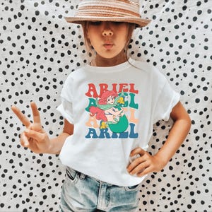 May include: A white t-shirt featuring a retro-style graphic of Ariel the Little Mermaid. The graphic depicts Ariel with her iconic red hair and green tail, holding a yellow fish. The text "ARIEL" is repeated three times in a rainbow of colours.