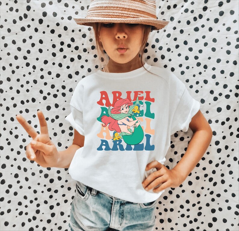 May include: A white t-shirt featuring a retro-style graphic of Ariel the Little Mermaid. The graphic depicts Ariel with her iconic red hair and green tail, holding a yellow fish. The text "ARIEL" is repeated three times in a rainbow of colours.