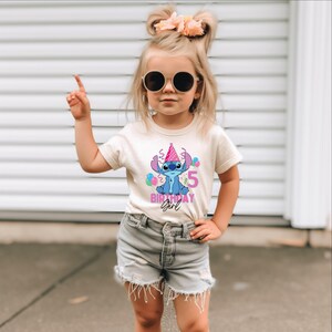 May include: A beige t-shirt with a blue cartoon character wearing a pink party hat and the number 5. The text "BIRTHDAY GIRL" is below the character. The shirt is worn by a young child.