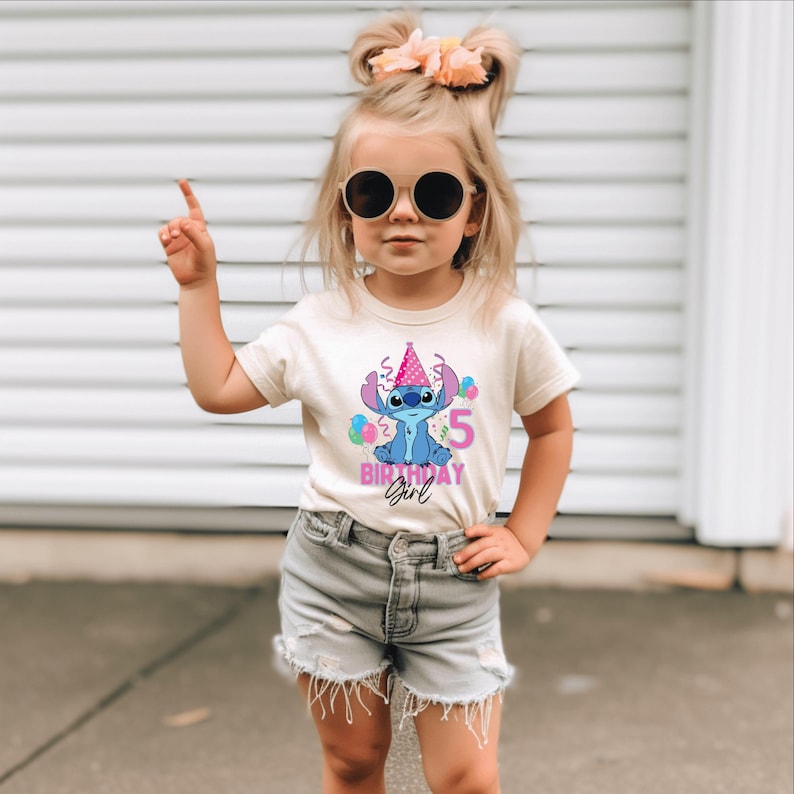 May include: A beige t-shirt with a blue cartoon character wearing a pink party hat and the number 5. The text "BIRTHDAY GIRL" is below the character. The shirt is worn by a young child.