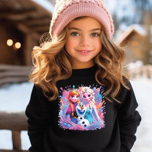 May include: A young girl wearing a black sweatshirt featuring a colorful graphic of Anna, Elsa, and Olaf from the movie Frozen. She is also wearing a pink knit beanie with a pom-pom. The background shows a snowy outdoor scene.