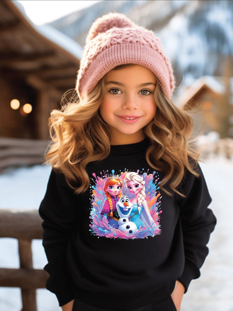 May include: A young girl wearing a black sweatshirt featuring a colorful graphic of Anna, Elsa, and Olaf from the movie Frozen. She is also wearing a pink knit beanie with a pom-pom. The background shows a snowy outdoor scene.