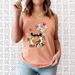May include: Peach-colored tank top featuring a cartoon graphic of Chip and Dale holding hands, with colorful Mickey Mouse balloons. The design includes popcorn, and other festive elements. The top is made of a soft material.