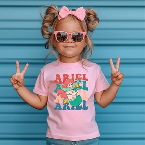 May include: A young girl wearing pink sunglasses and a pink T-shirt with a graphic of Ariel the mermaid from The Little Mermaid. The shirt has the text "Ariel" repeated in a retro font.