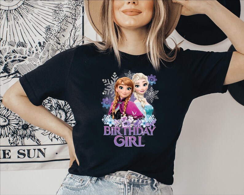 K&ouml;nnte beinhalten: Schwarzes T-Shirt mit einer Grafik von Anna und Elsa, animierten Prinzessinnen, umgeben von Schneeflocken und Blumen. Der Text "BIRTHDAY GIRL" steht darunter in Lila. Das T-Shirt wird von einer Person getragen.