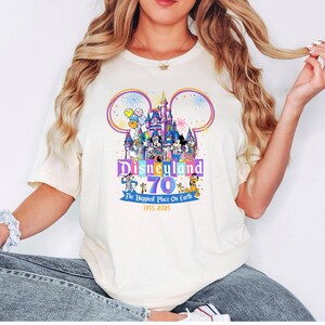 May include: Cream-colored t-shirt featuring a colorful Disneyland 70th anniversary graphic. The design includes the Disneyland castle, Mickey Mouse ears, and various Disney characters. Text reads "Disneyland 70 The Happiest Place On Earth 1955-2025."