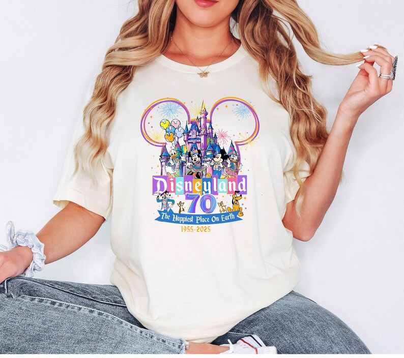 May include: Cream-colored t-shirt featuring a colorful Disneyland 70th anniversary graphic. The design includes the Disneyland castle, Mickey Mouse ears, and various Disney characters. Text reads "Disneyland 70 The Happiest Place On Earth 1955-2025."
